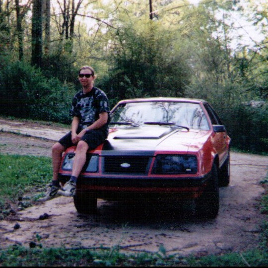 A younger me wearing sunglasses, sitting on the hood of my red Mustang
